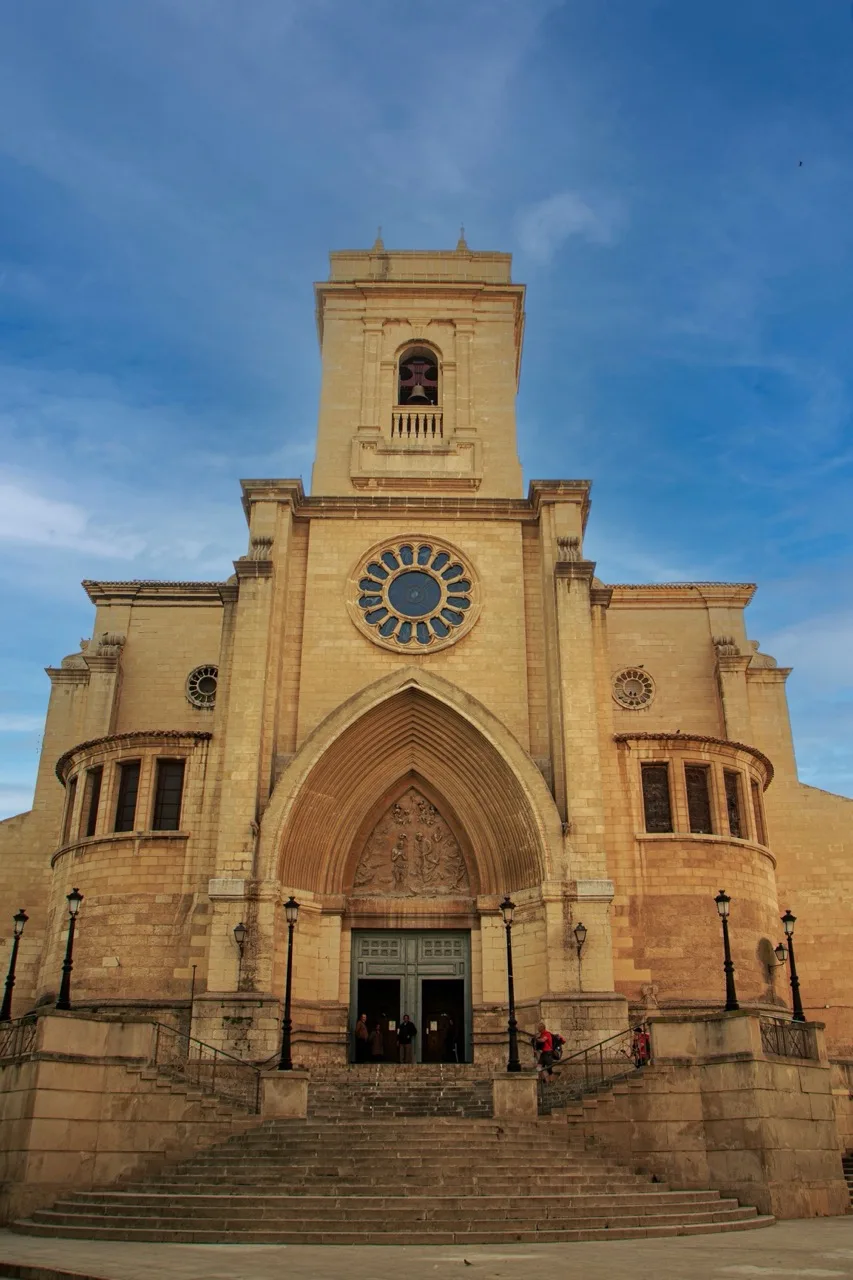 Se ve la fachada de la Catedral de Albacete, con torre central, gran rosetón, arco apuntado y escalinata de piedra en primer plano, bajo un cielo azul.