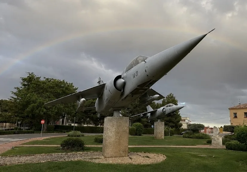 2 cazas Mirage F1 grises se alzan sobre un pedestal en una plazoleta, bajo un arcoíris tenue y un cielo nublado en Albacete, con árboles y césped verde alrededor.