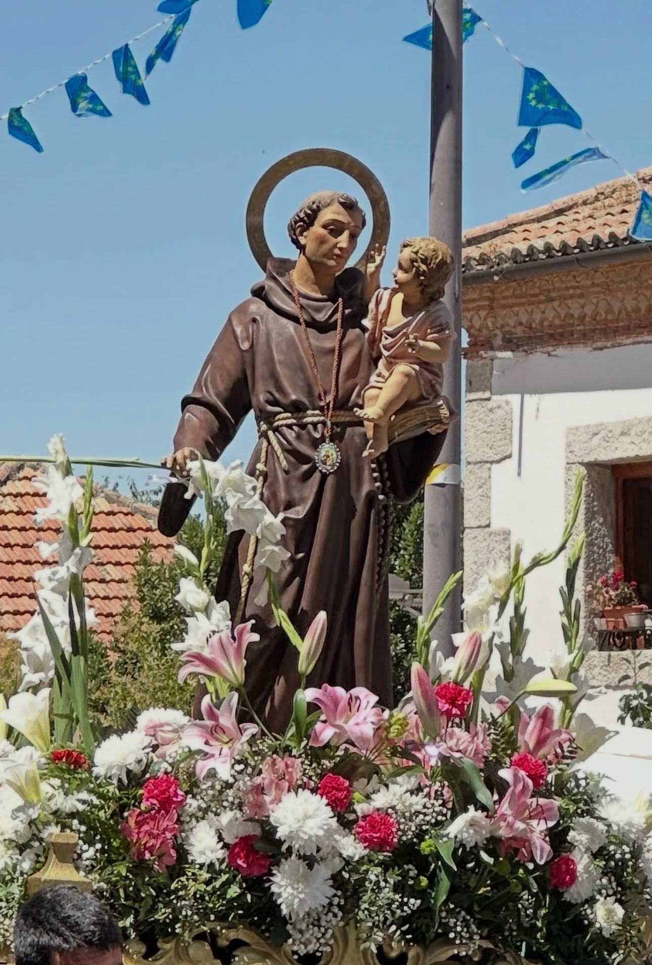 Durante una celebración al aire libre, la estatua de San Antonio de Padua sostiene al Niño Jesús entre flores blancas y rosas y banderas azules.