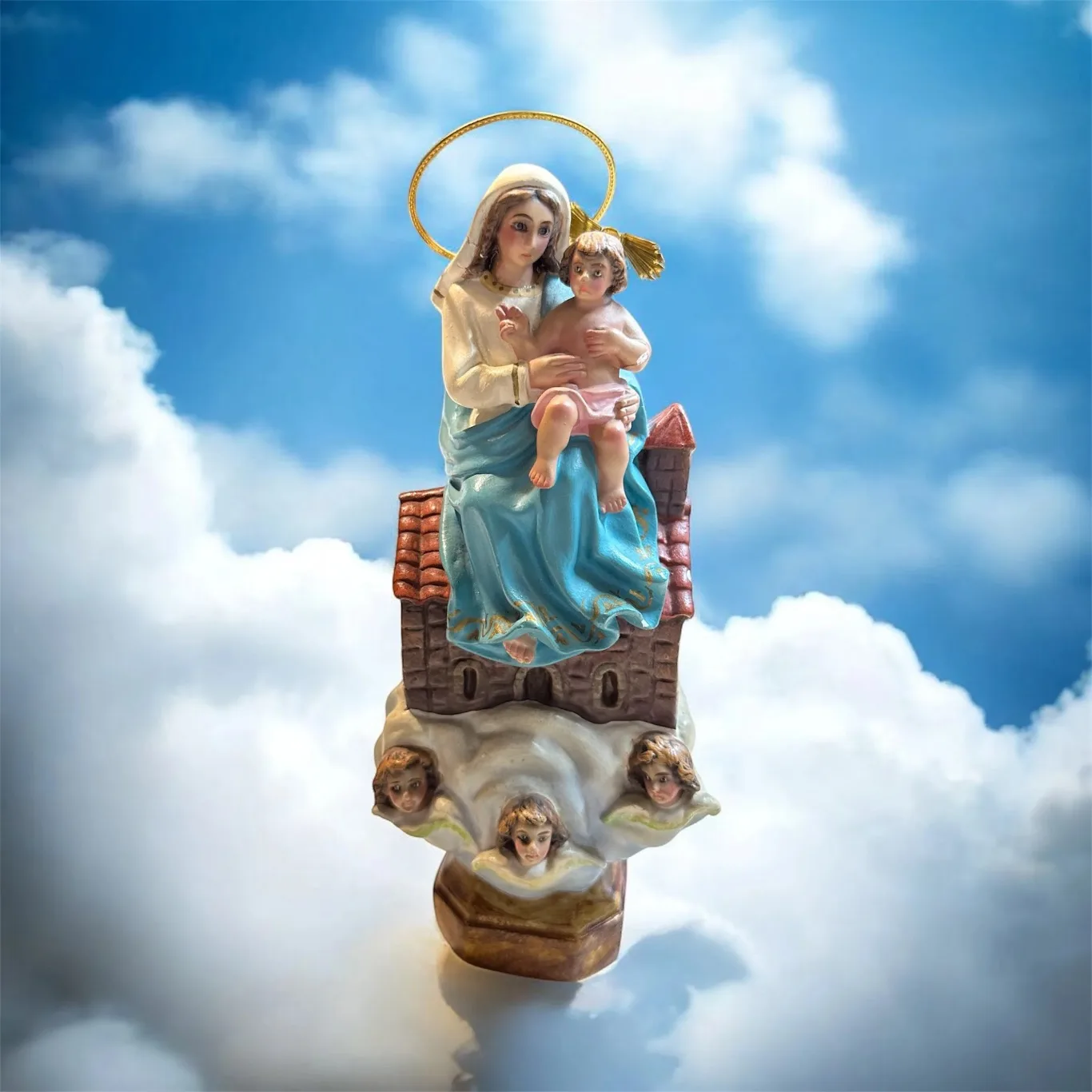Representación de la Virgen de Loreto con el Niño Jesús, coronada y sentada sobre nubes, frente a un cielo azul con luz suave y pequeñas casas.